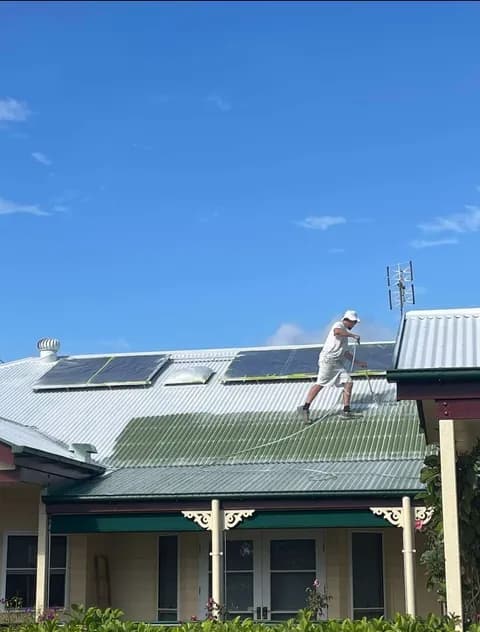 Worker in white spraying green paint onto a corrugated metal roof under blue skies.